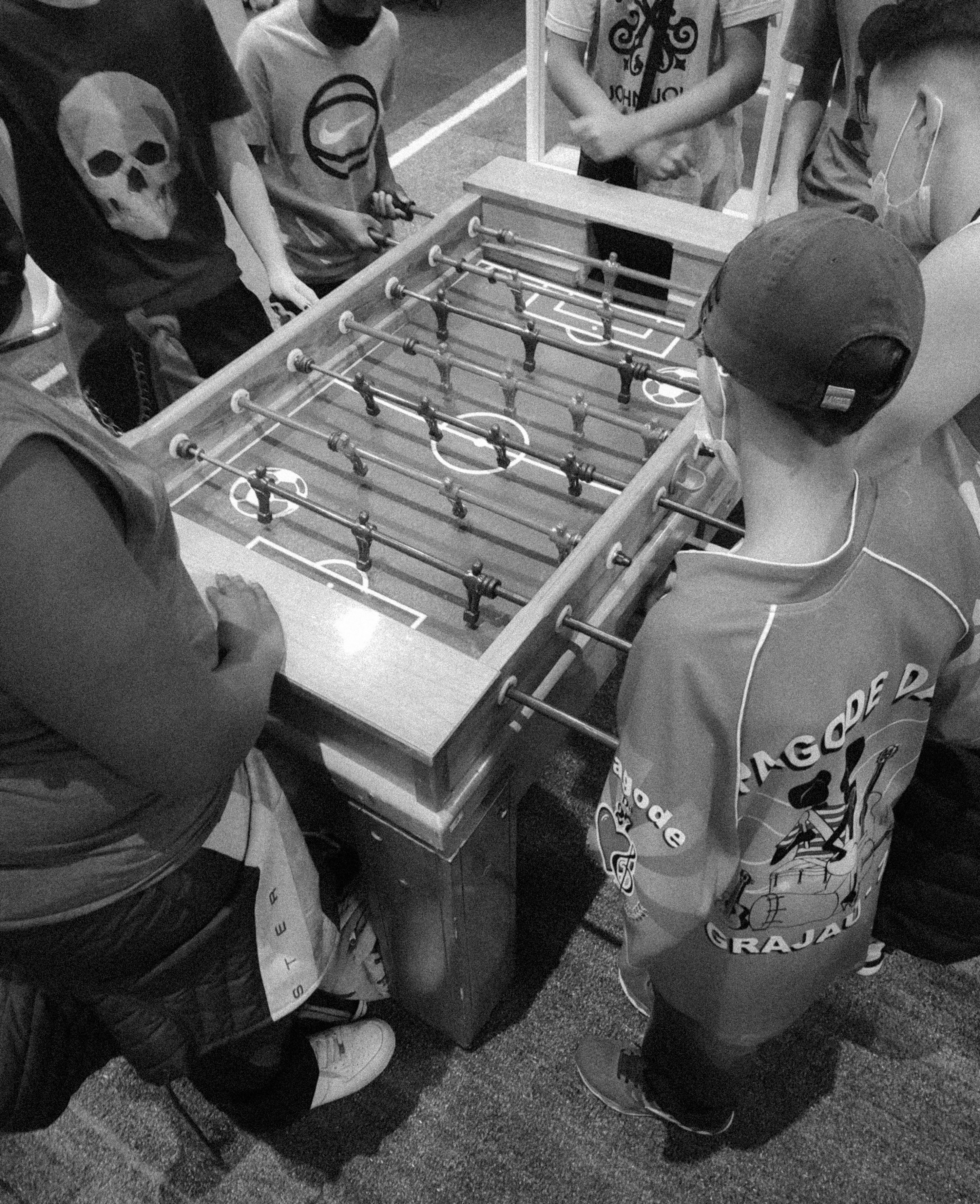 uma foto em preto e branco tirada de cima que mostra uma mesa de pebolim e várias pessoas em volta